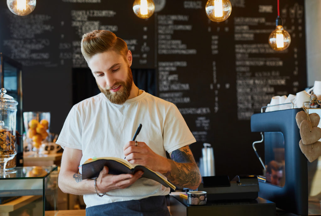 Smiling barista tracks decreasing business expenses in notebook