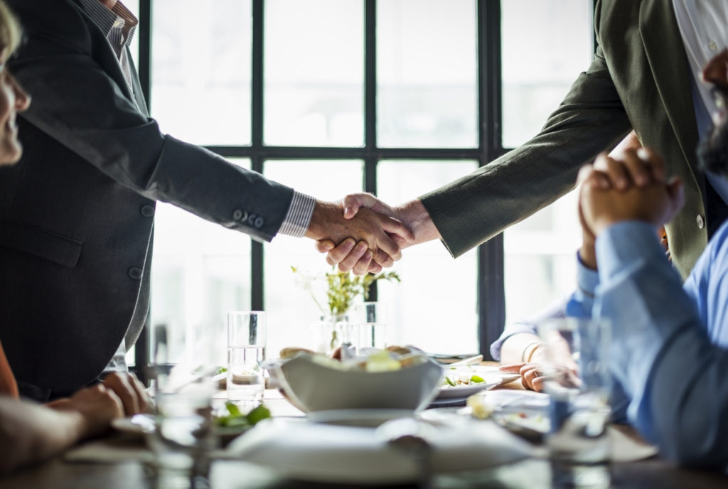Two men shake hands over a meal, closing a transfer of ownership deal
